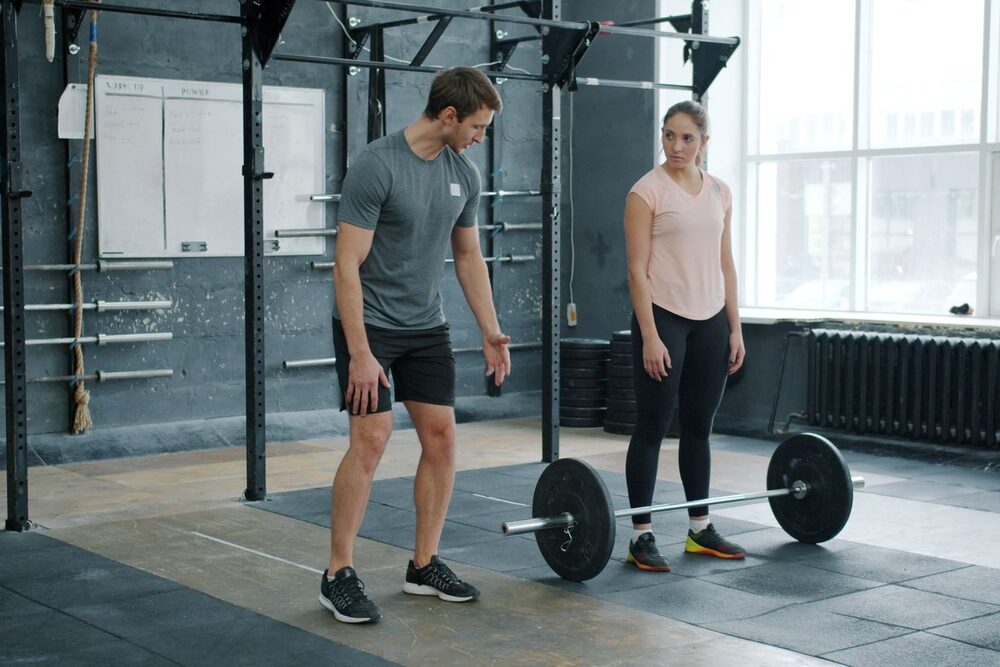 A personal trainer coaching a client on barbell technique in a bright gym