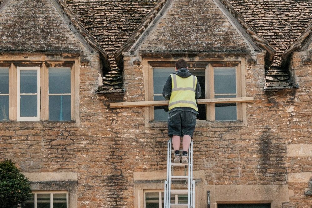 A tradesperson in a hi-vis vest up a ladder repairing the front of a stone house